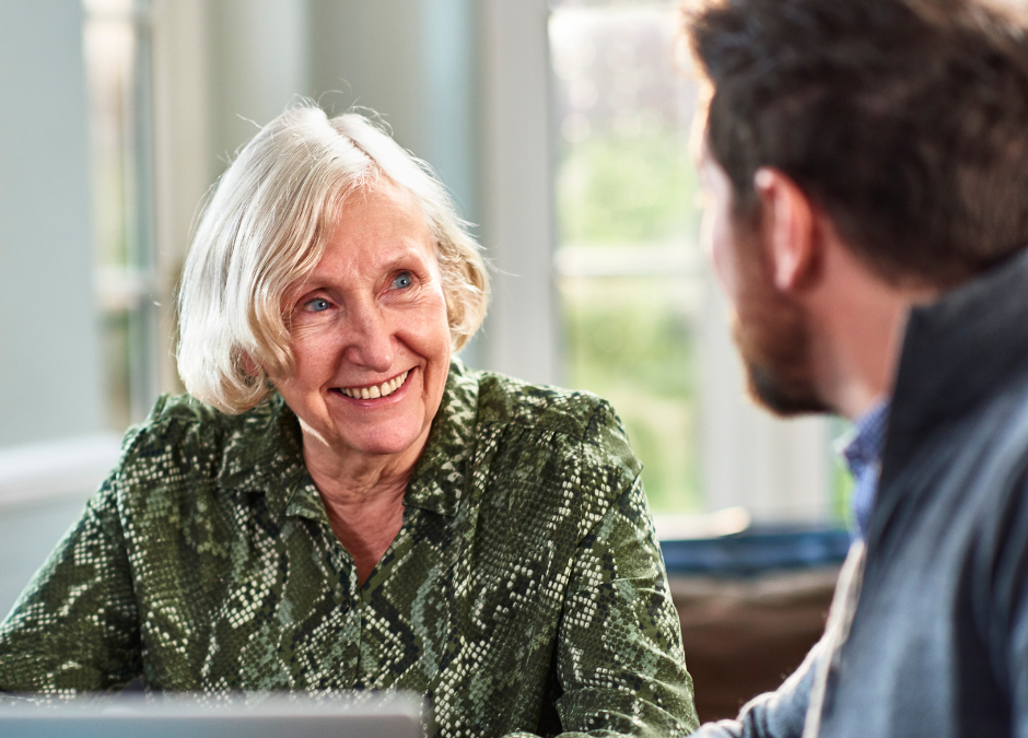 Older woman smiles talking to man with beard who we see side on with laptop edge in foreground